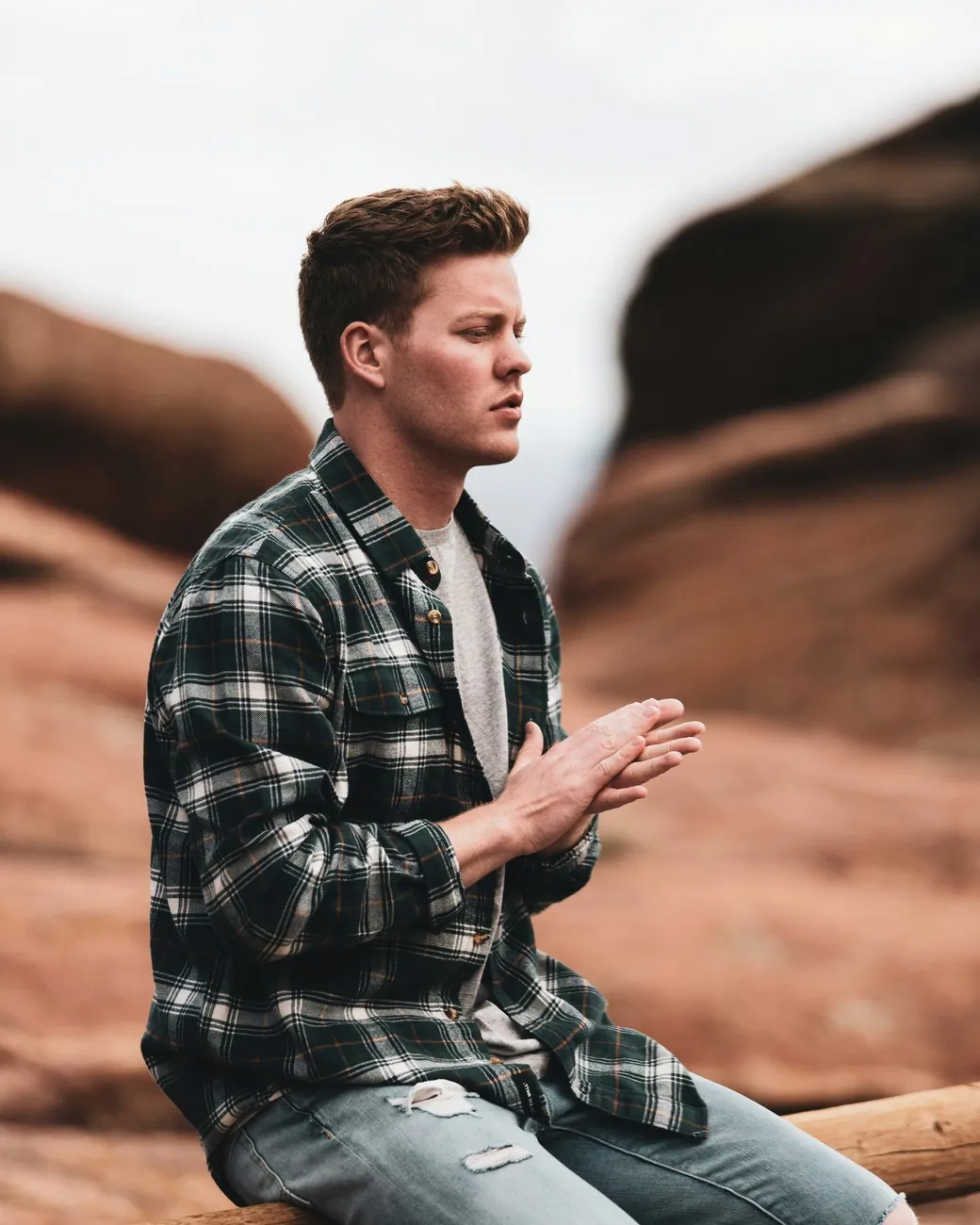 A young man in a plaid shirt sits on a wooden rail in front of red rock, hands pressed together, eyes down.